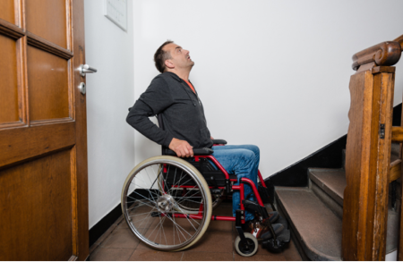 Man in rolstoel staat onderaan de trap van een woning en kijkt omhoog