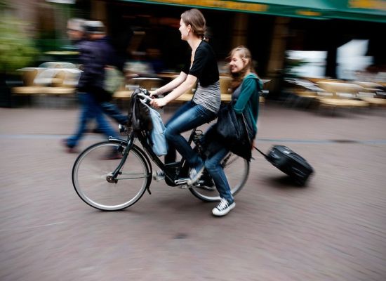 Twee jongedames op de fiets. Een achterop met een koffer op wieltjes