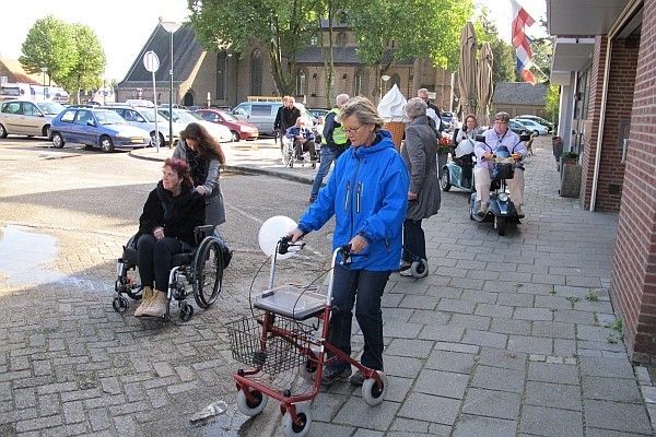 Gezelschap met rollators, rolstoelen, scootmobielen en blindegeleidestokken op weg naar het gemeentehuis. Obstakel hoge stoeprand.