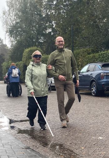 Twee personen lopen op straat. De een loopt met een blindegeleidestok en oogmasker op. De ander geeft een arm ter ondersteuning.