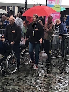 een groep wandelaars inclusief mensen in rolstoel met duwer, lopend in de regen met paraplu's