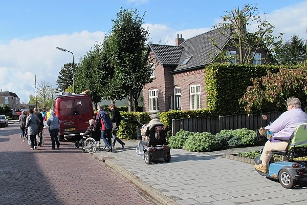 Gezelschap met rollators, rolstoelen, scootmobielen en blindenstokken bij op stoep geparkeerde bus