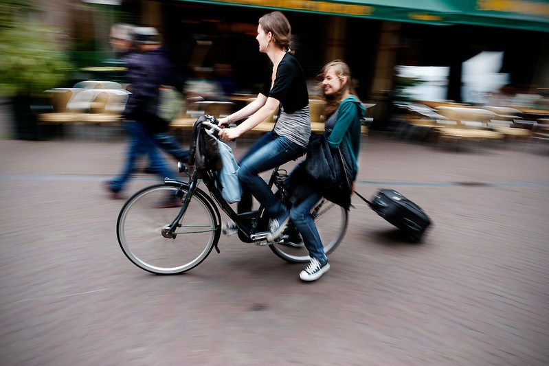 Twee jongedames op de fiets. Een achterop met een koffer op wieltjes