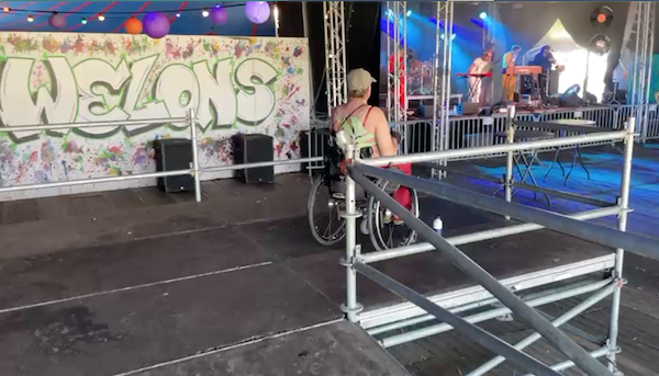 Verhoogd uitkijkdek/podium met vrouw in rolstoel kijkend en luisterend naar het optreden van een band