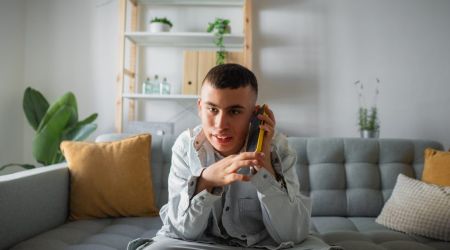 Jongeman is aan het bellen met zijn mobieltje, zittend op de bank in zijn woonkamer