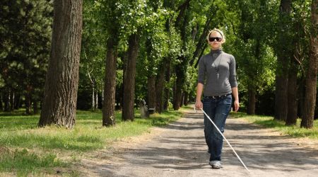 Een vrouw met blindegeleidestok en zonnebril op loopt in de bossen