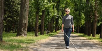Een vrouw met blindegeleidestok en zonnebril op loopt in de bossen